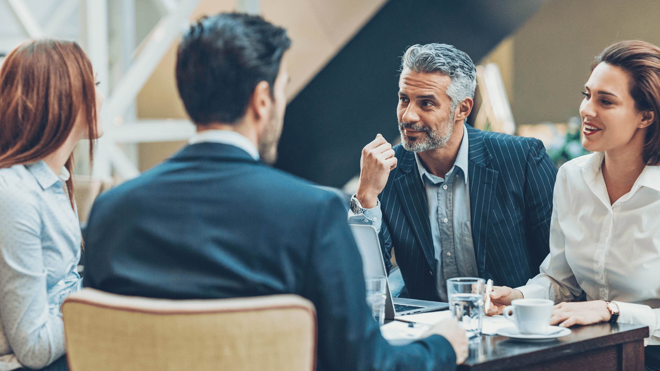 Four people in business attire having a discussion around a table with laptops and coffee cups in a modern office setting.