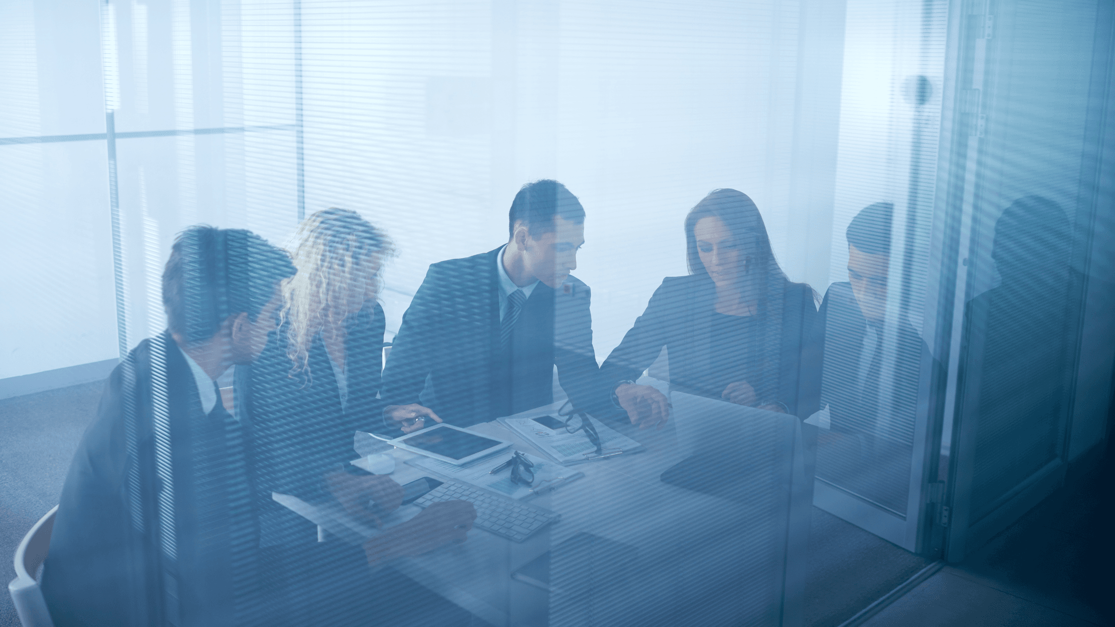 Business meeting with five people in formal attire around a table, viewed through frosted glass, discussing strategic plans.