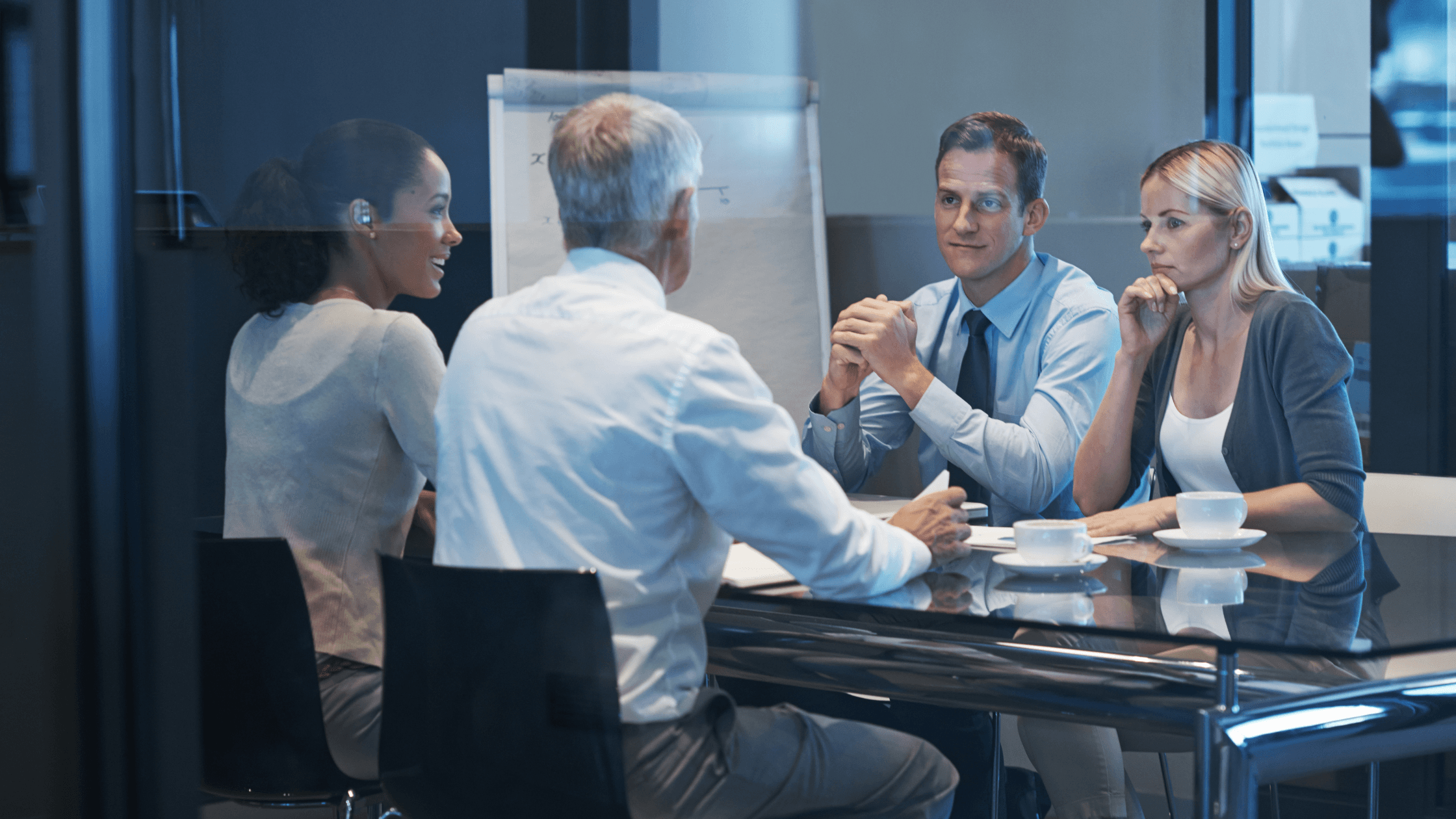 Four people in a business meeting around a glass table, with a flip chart in the background and coffee cups on the table.
