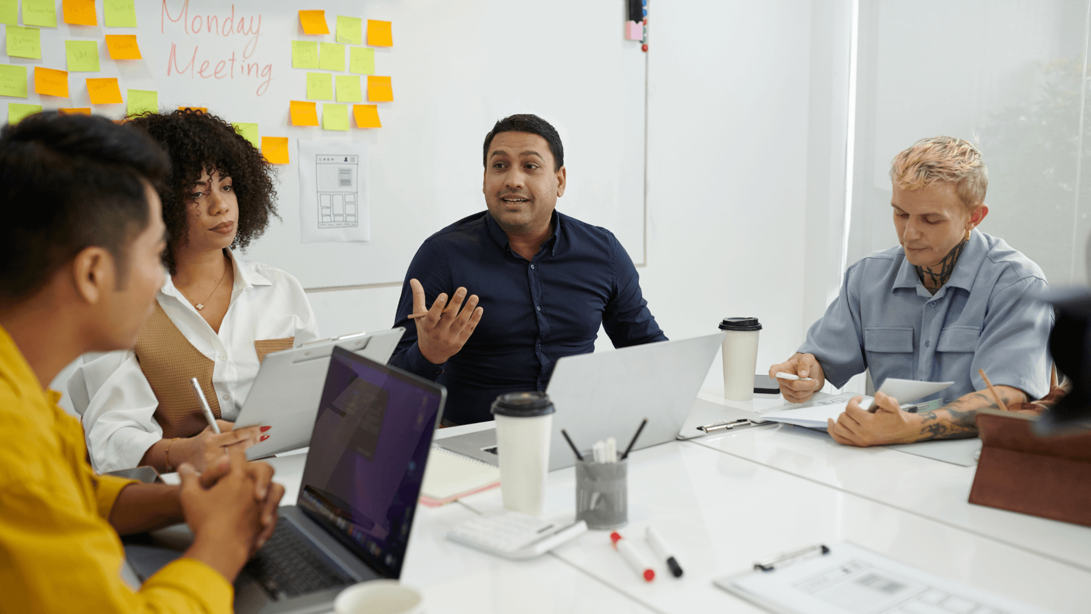 A group of four colleagues in a meeting room with laptops and coffee cups, discussing, while brainstorm notes are on a whiteboard behind them.
