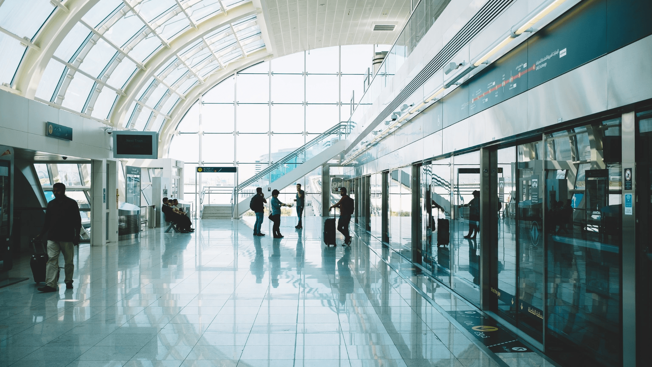 Spacious airport terminal with large windows, travelers walking, and an escalator leading to another level. Bright natural light fills the space.