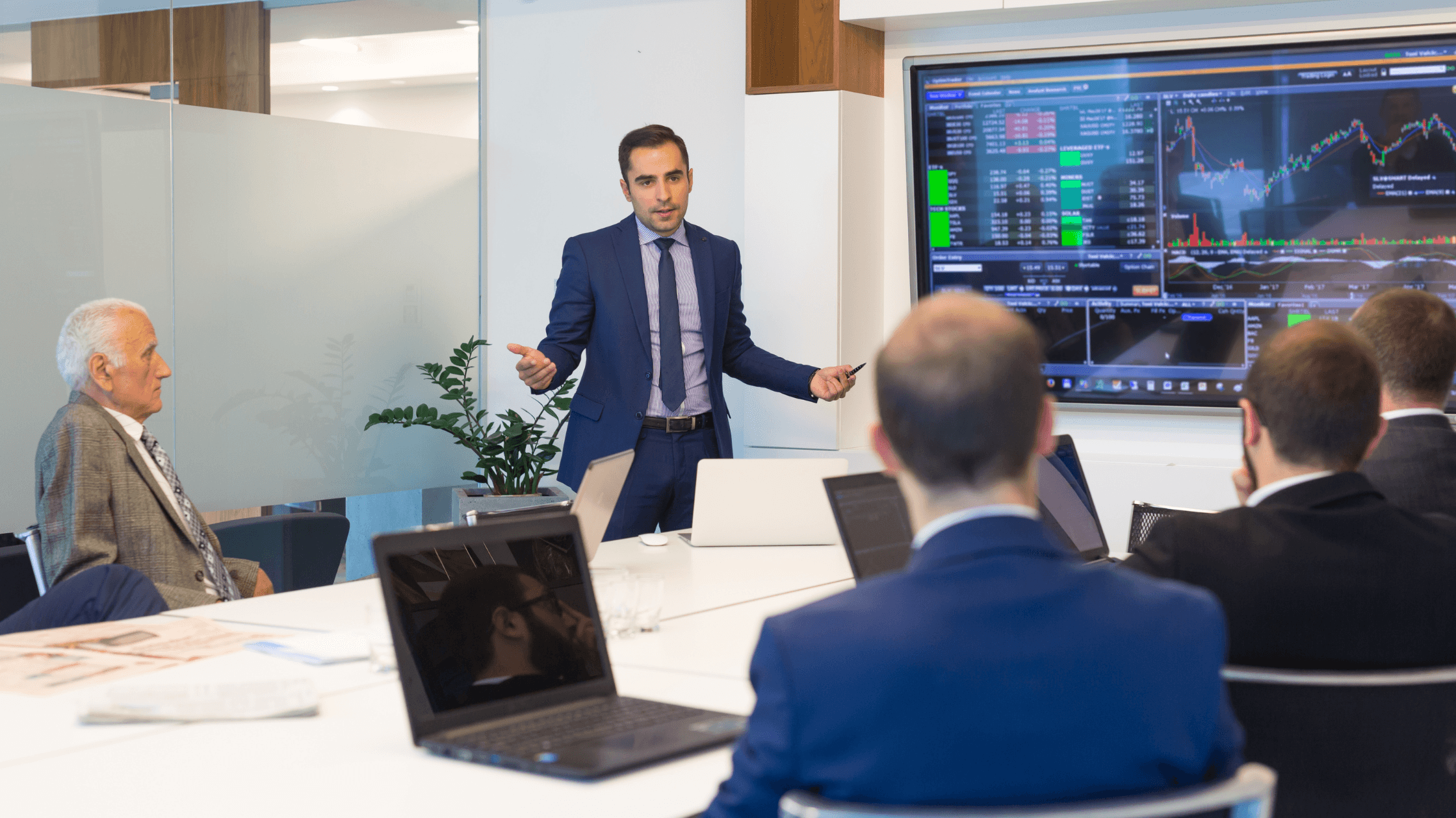 Businessman in a suit presenting stock market data on a large screen to a group of colleagues in a conference room.