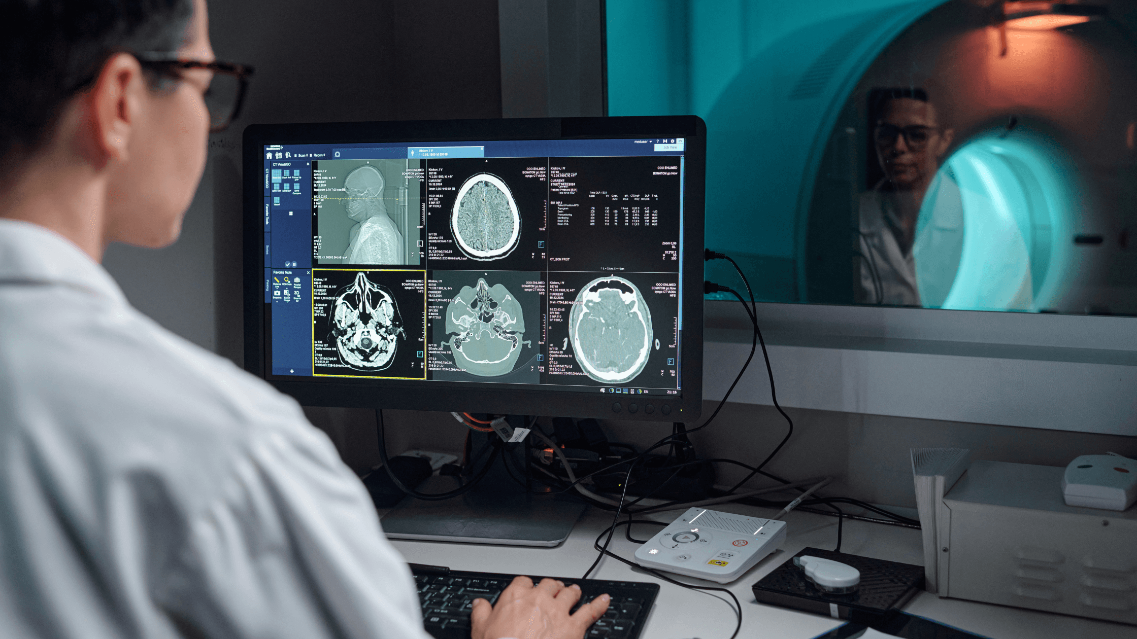 Technician reviews brain scan images on a computer monitor, while an MRI machine is visible through a window in the background.