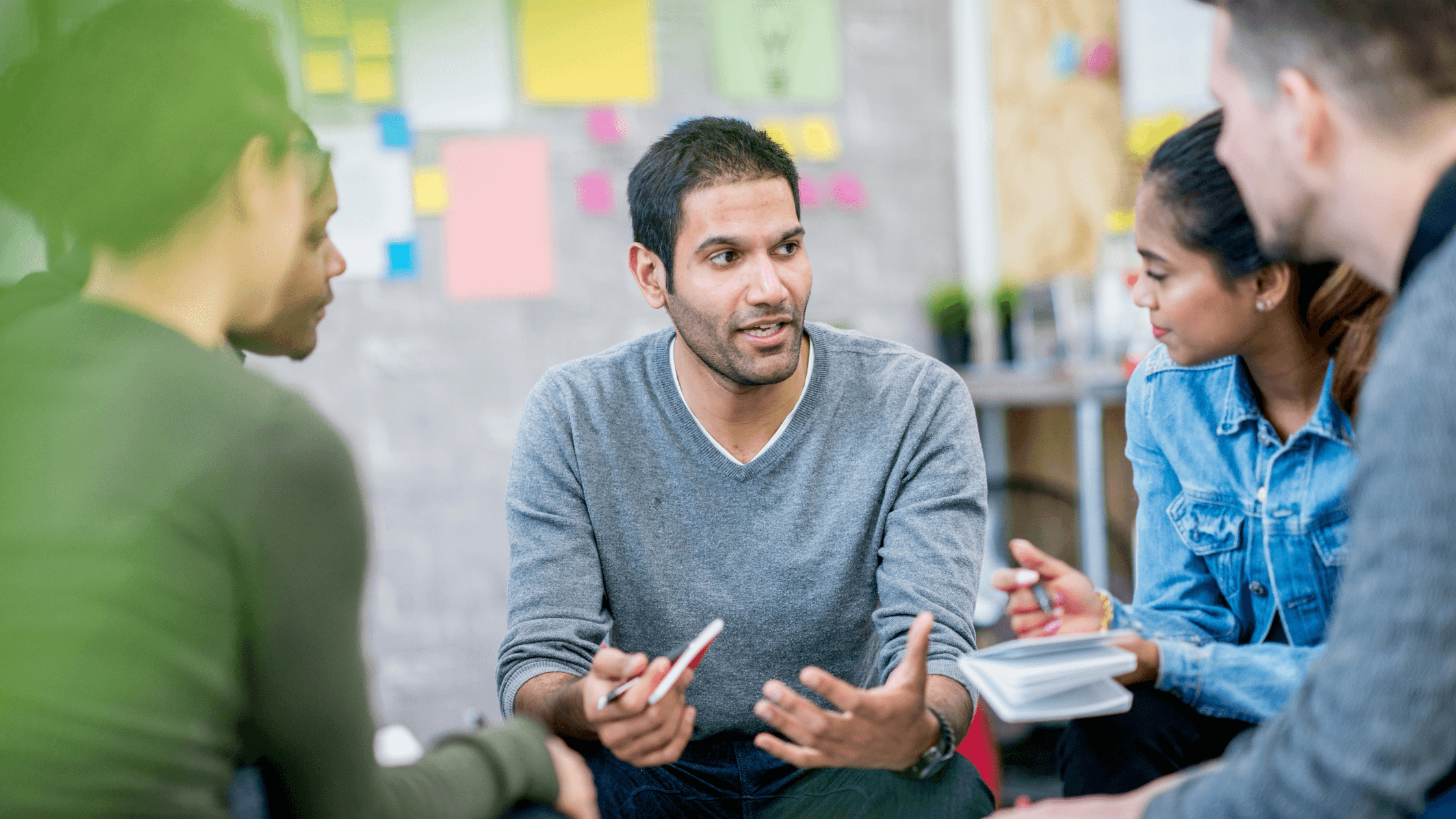 A group of five people sitting in a circle, discussing and taking notes in a scenario planning meeting with colorful sticky notes on the wall.