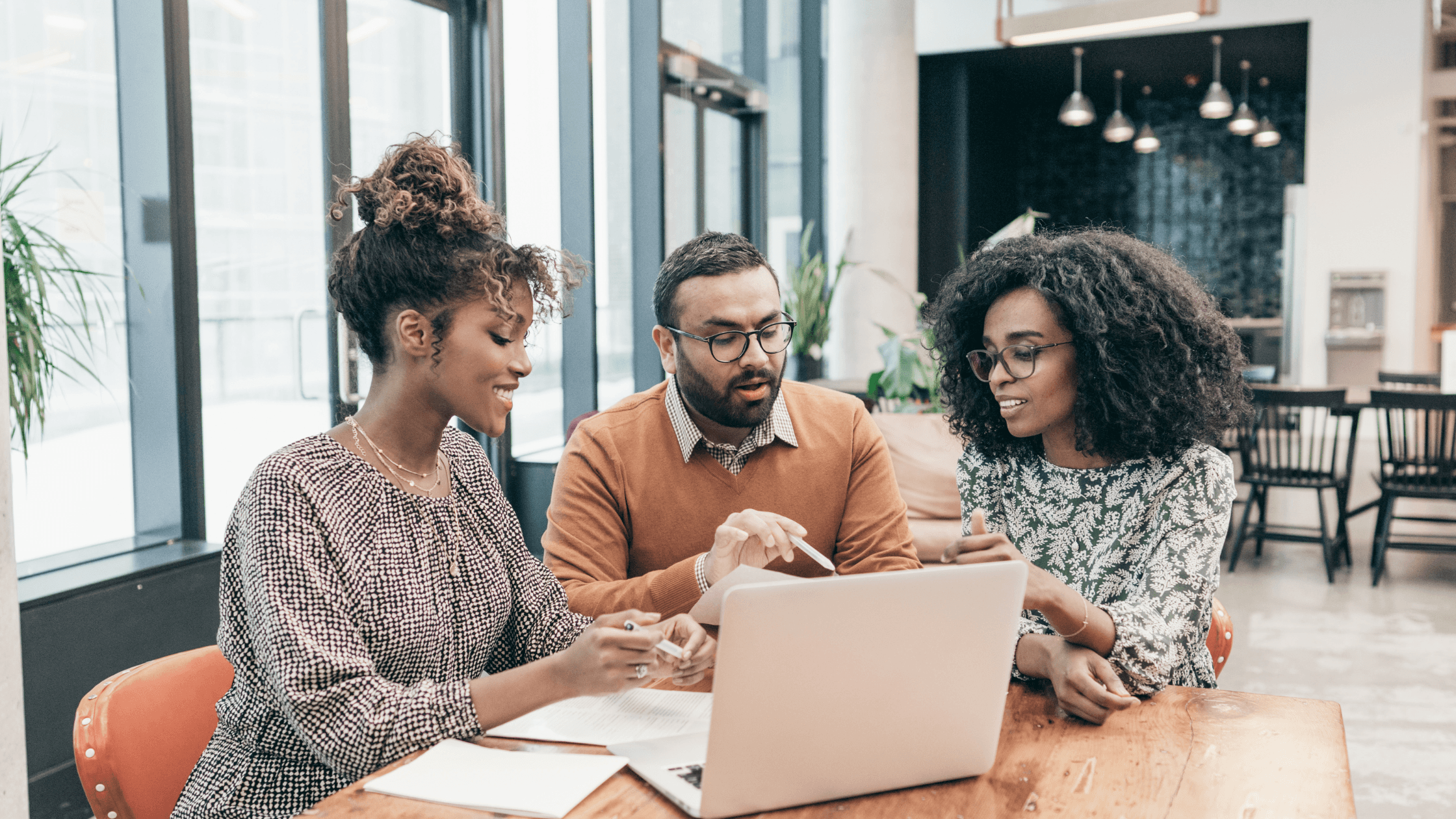 Three colleagues discussing work at a table with a laptop, in a modern office with large windows and plants.