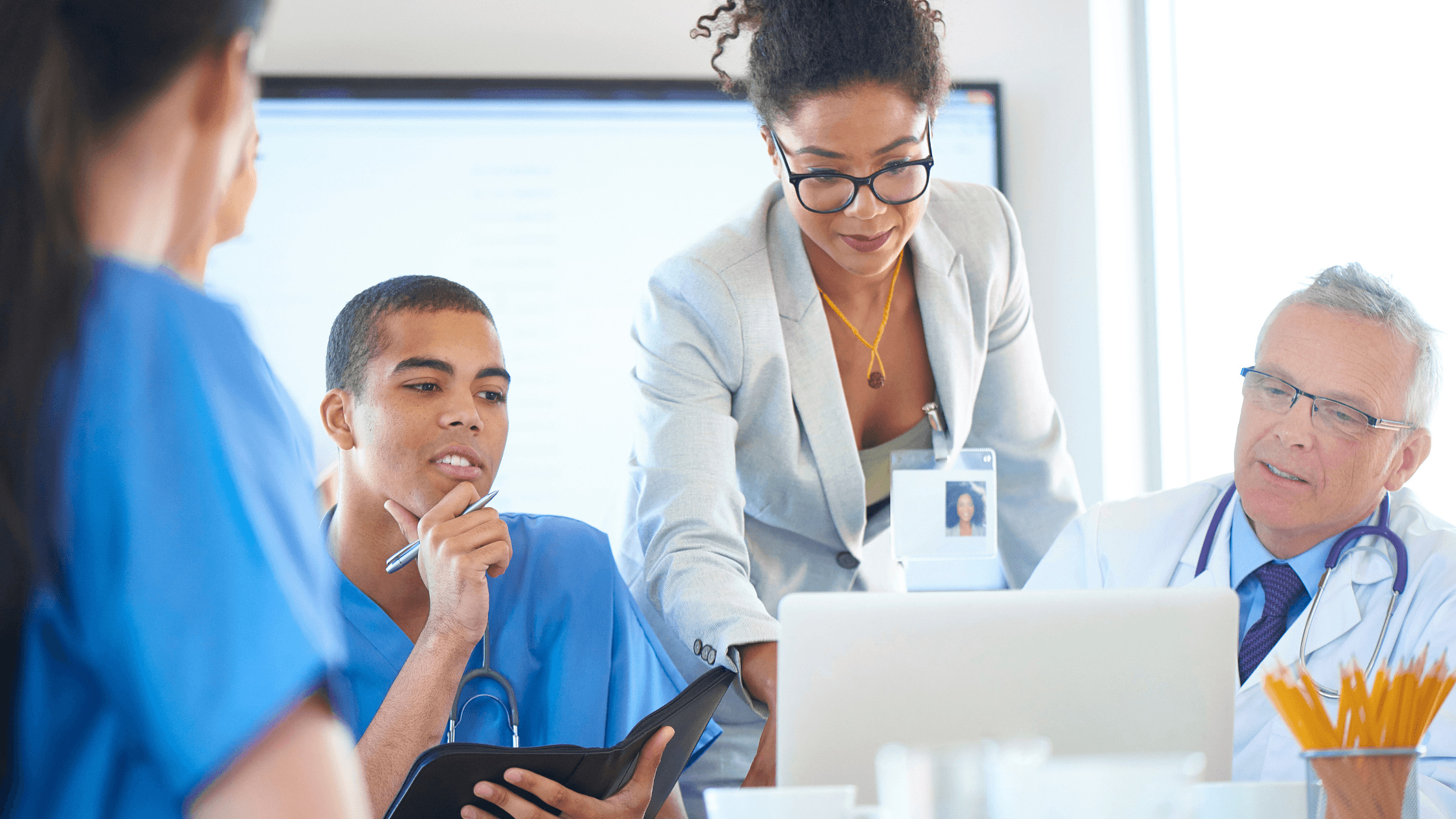 Medical team in discussion; one member points to a laptop screen. Others are in blue scrubs, one in a white coat, engaged and attentive.