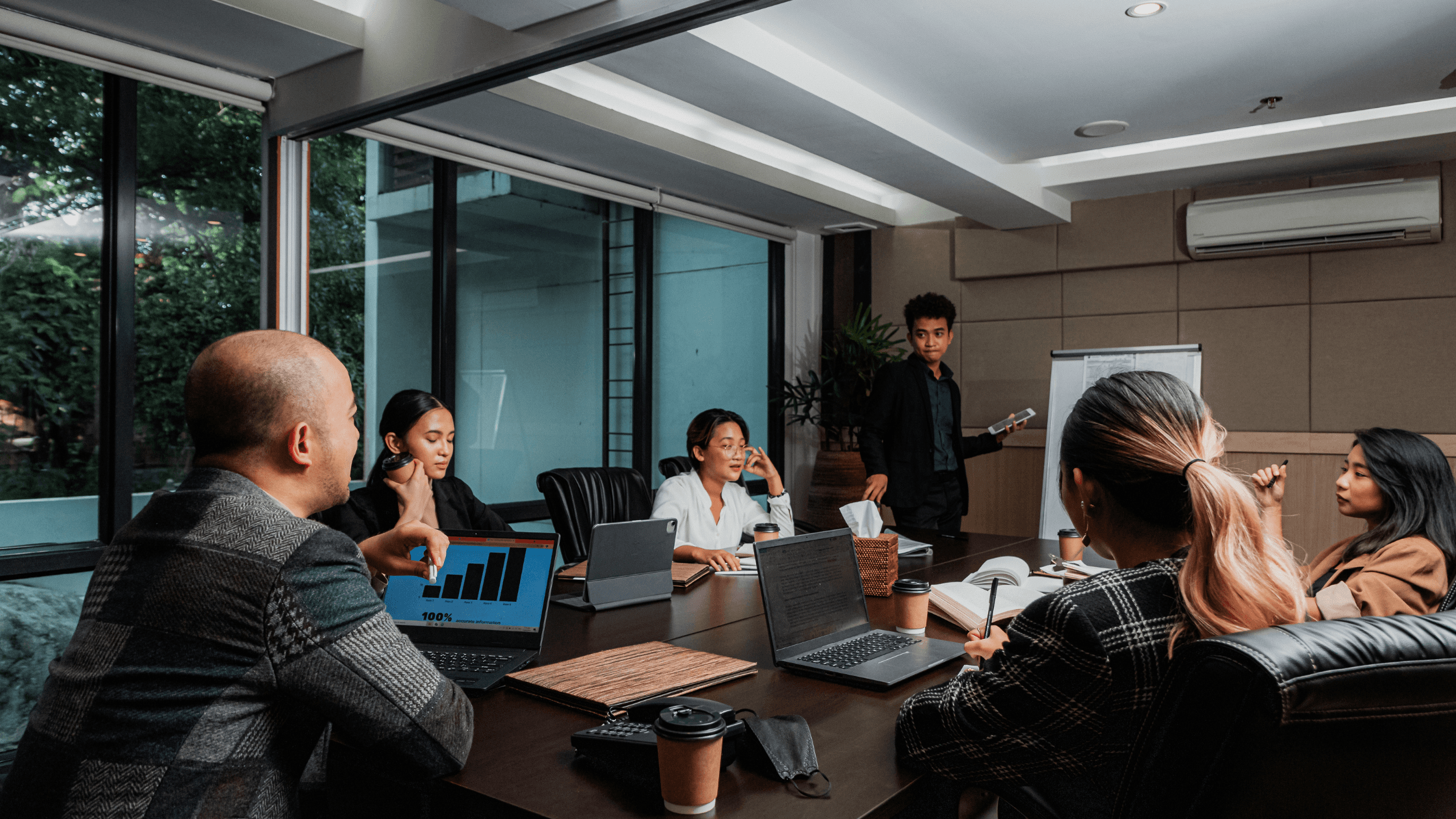 People in a modern conference room having a meeting; one person presents data on a laptop and a whiteboard.