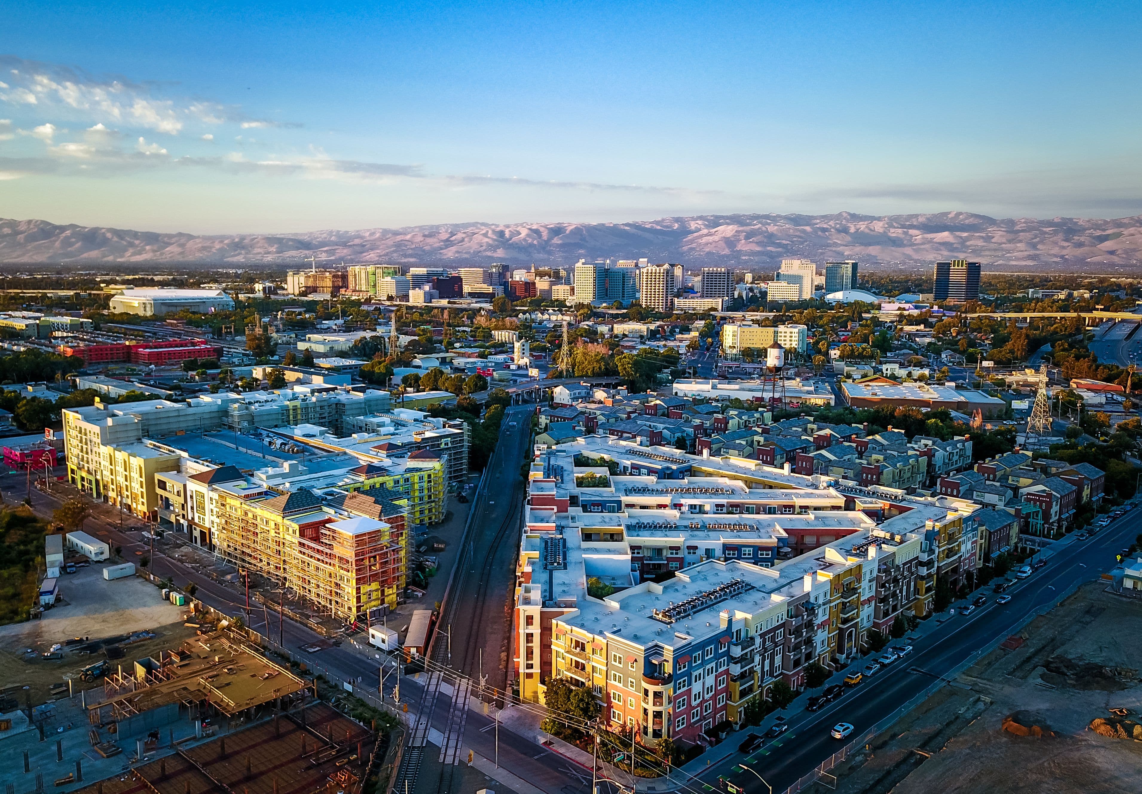 Aerial view of a cityscape with colorful buildings, roads, and distant mountains under a clear blue sky at sunset.