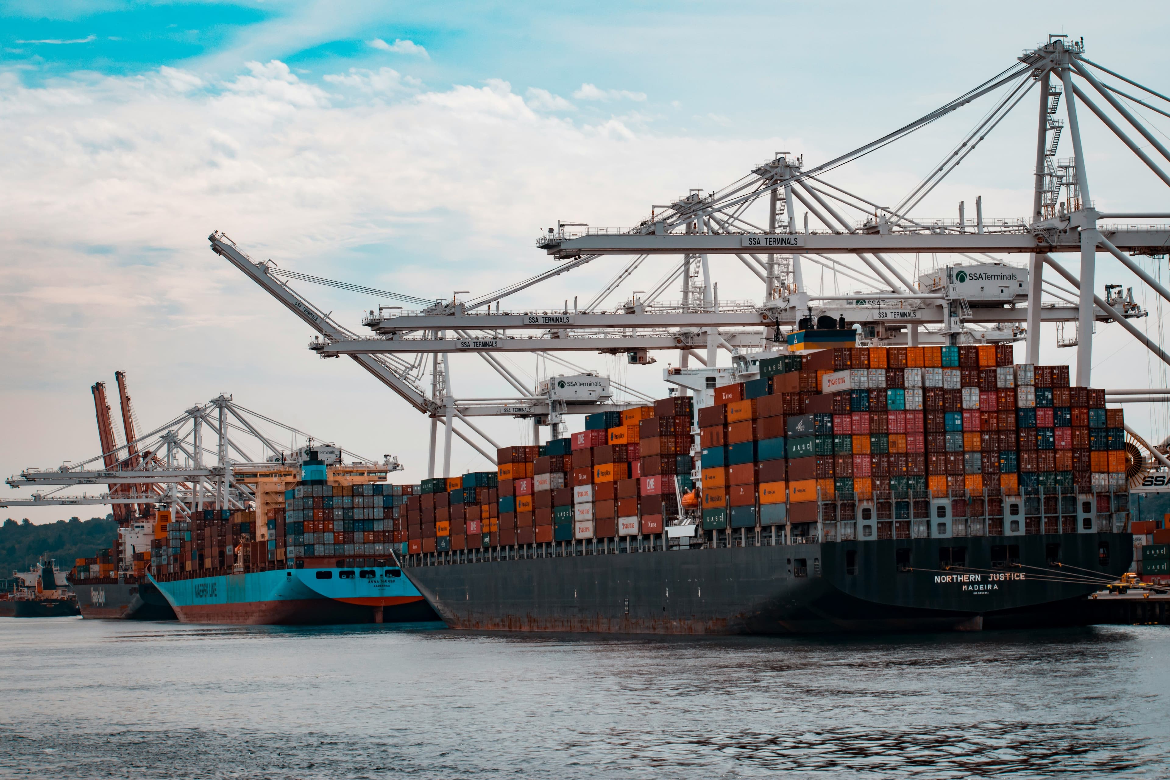 Cargo ships loaded with colorful containers docked at a busy port, with large cranes in the background under a partly cloudy sky.