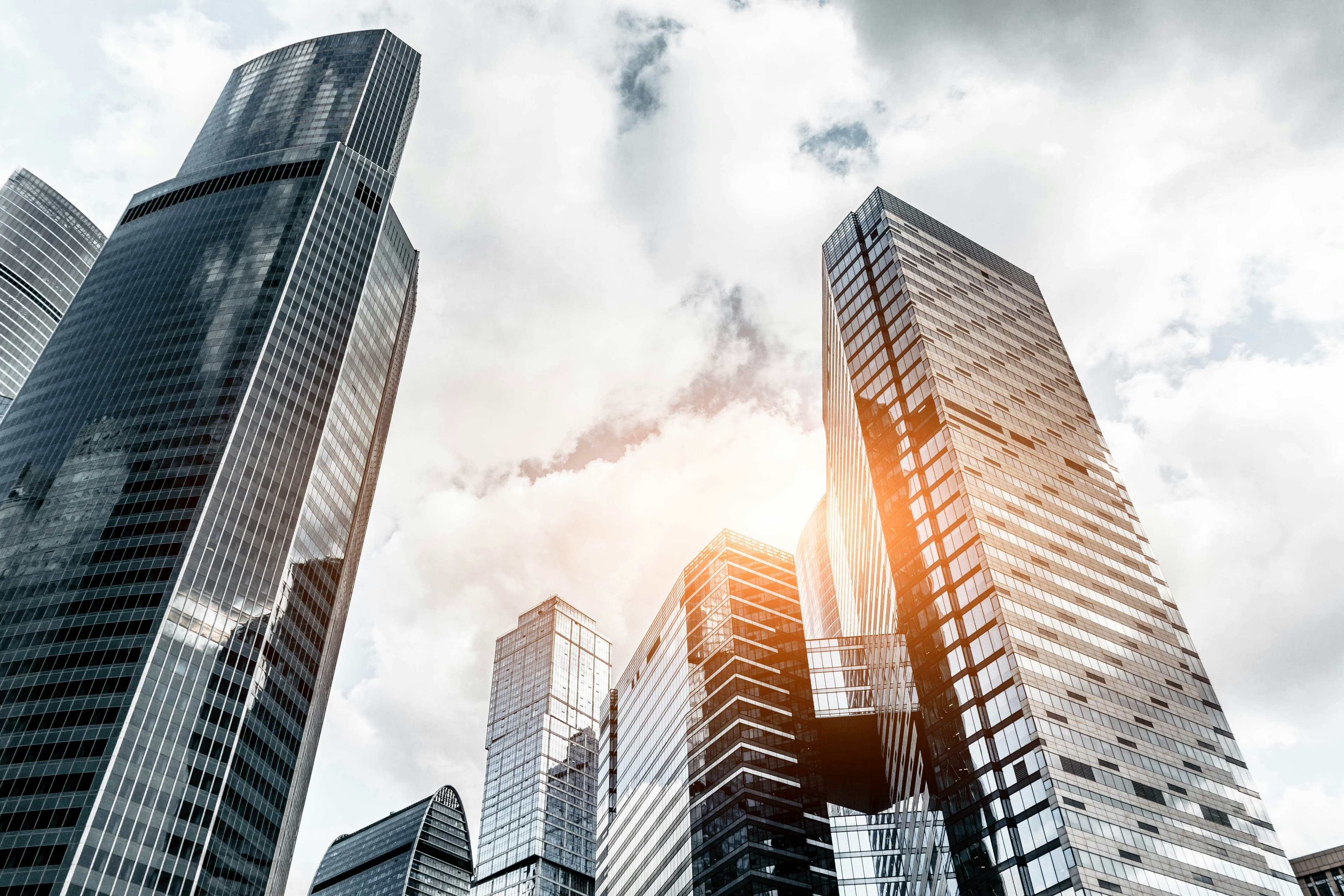 Tall modern skyscrapers with reflective glass facades against a cloudy sky, sunlight peeking through the buildings.