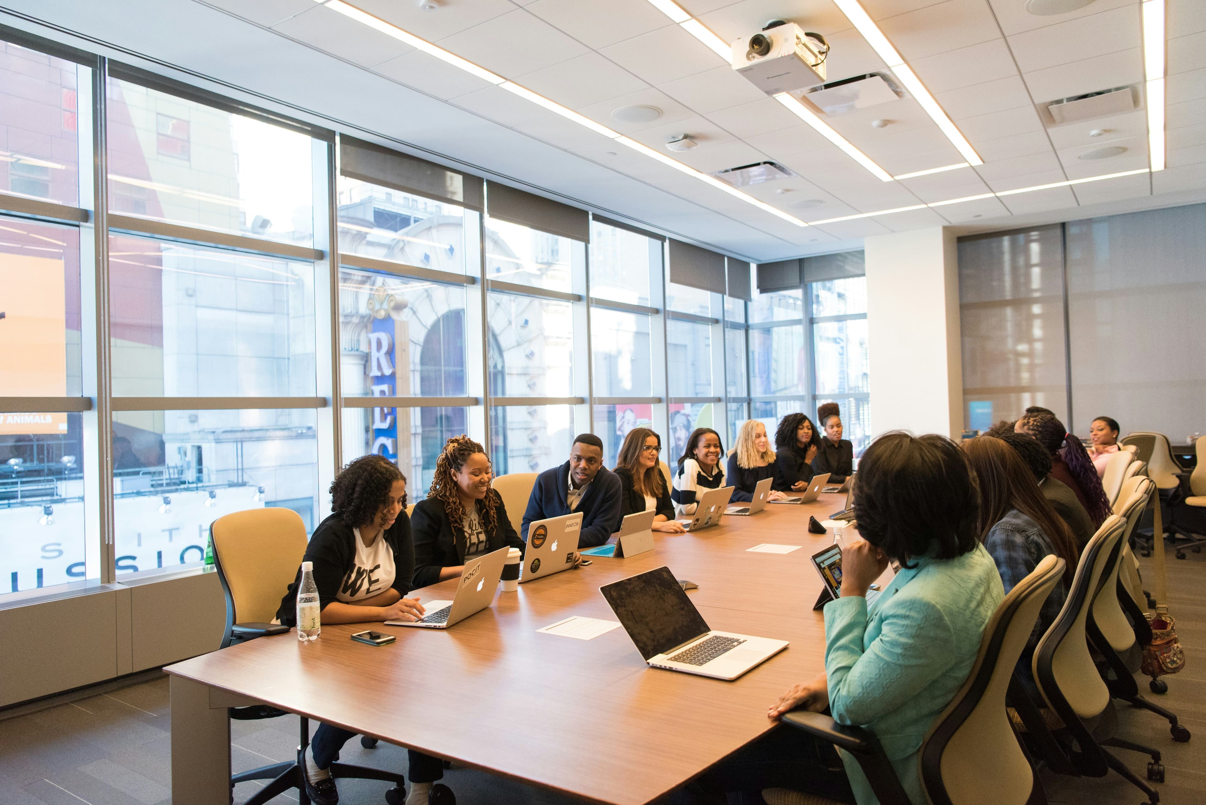 People sitting around a large conference table in a modern office, using laptops and engaging in discussion. Large windows in the background.