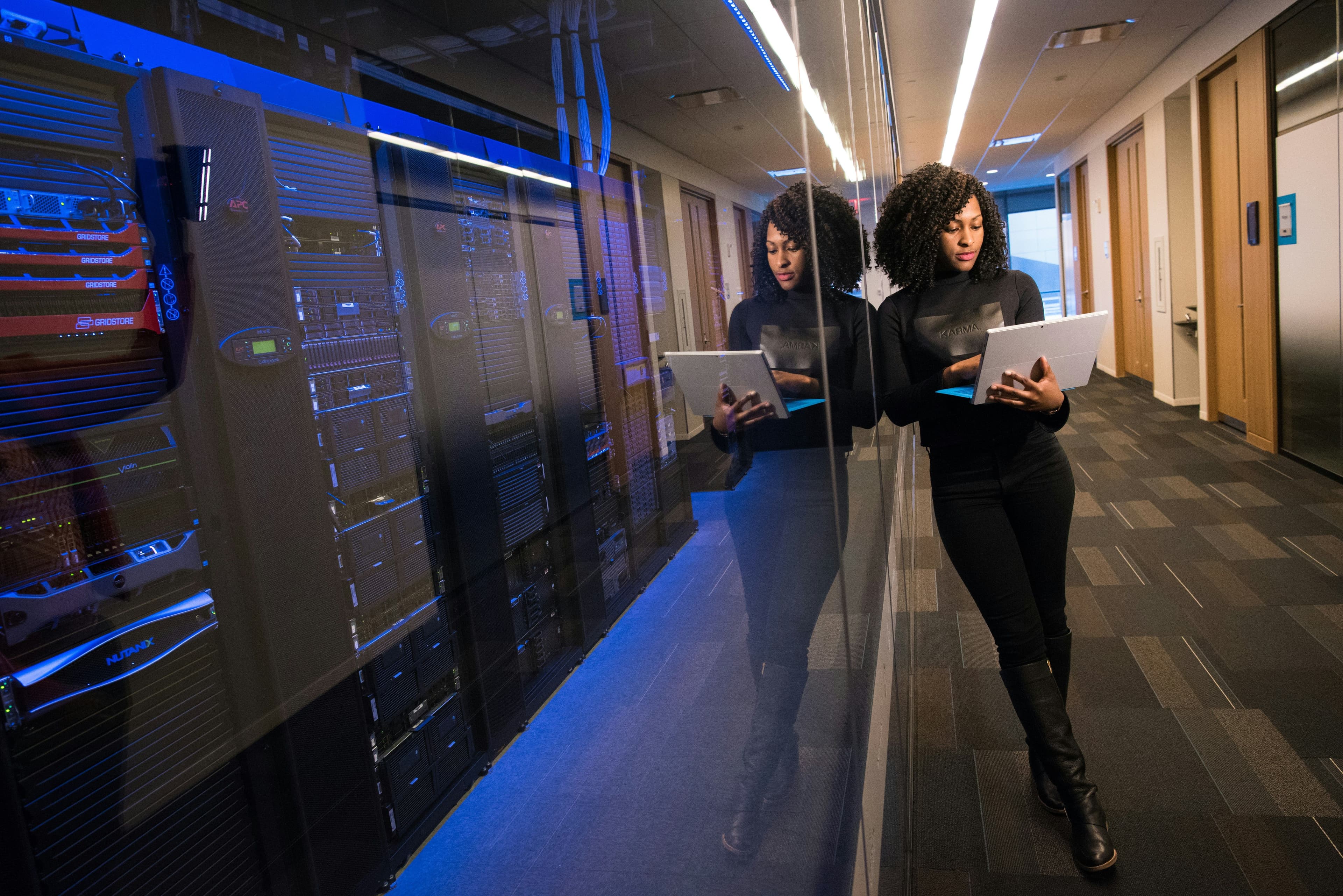 Woman leaning against a glass wall in a server room, holding a laptop, with blue lights reflecting on the glass.